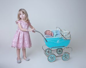 Proud older sister poses with baby brother in blue pram at their newborn photo session in Cambridge Ontario